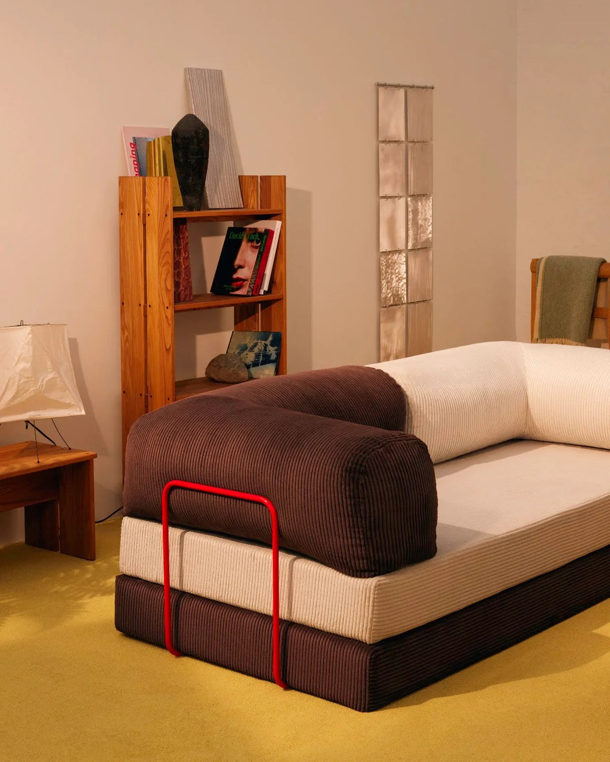 A deconstructed modular sofa displays layered cushions in cream and deep brown ribbed fabric with a striking red metal frame, staged in a warm Scandinavian showroom setting.