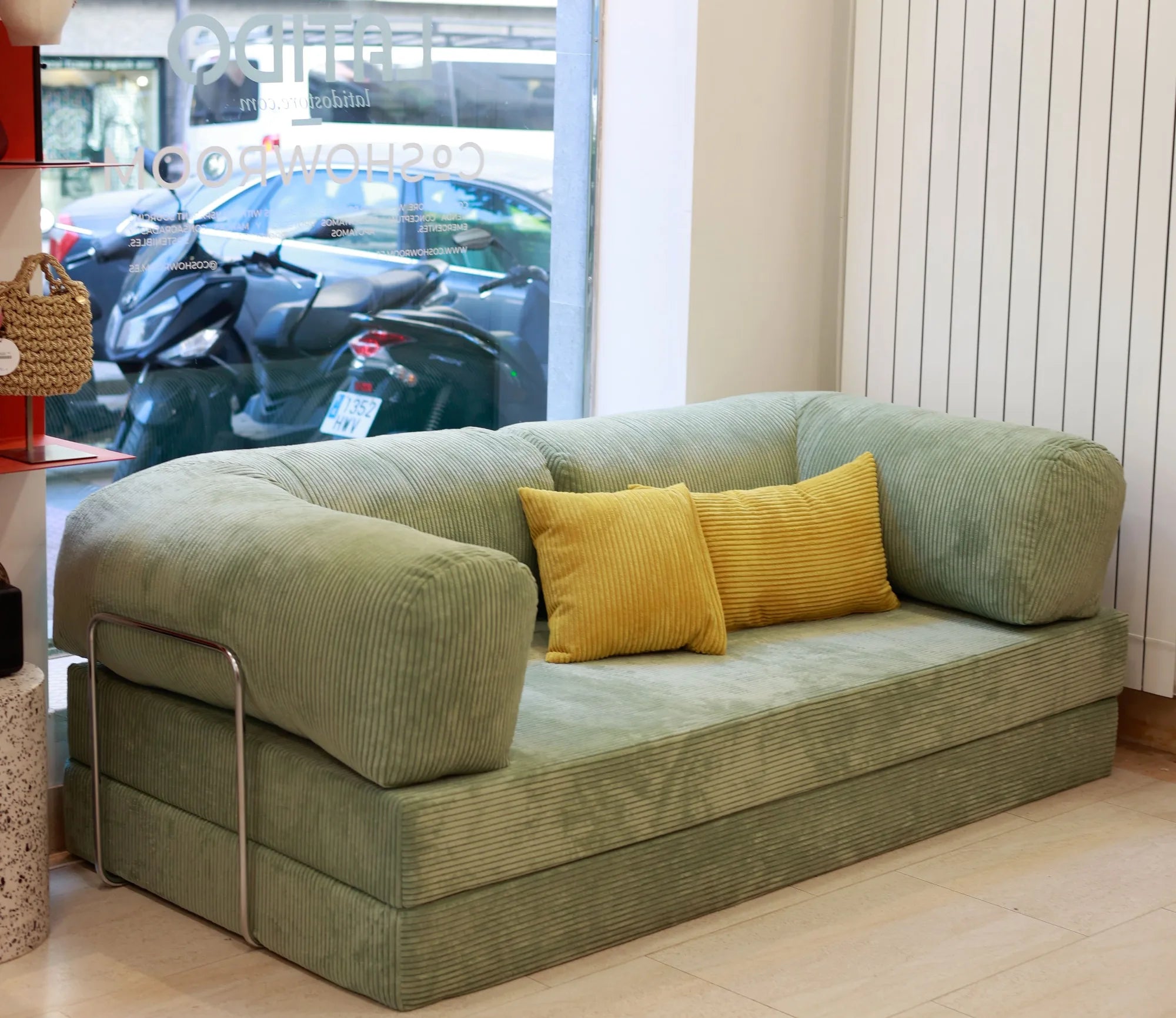 A contemporary sage green sectional sofa in corduroy with ribbed fabric upholstery, rounded rolled arms, and decorative mustard yellow cushions displayed in a bright showroom with street-facing window.